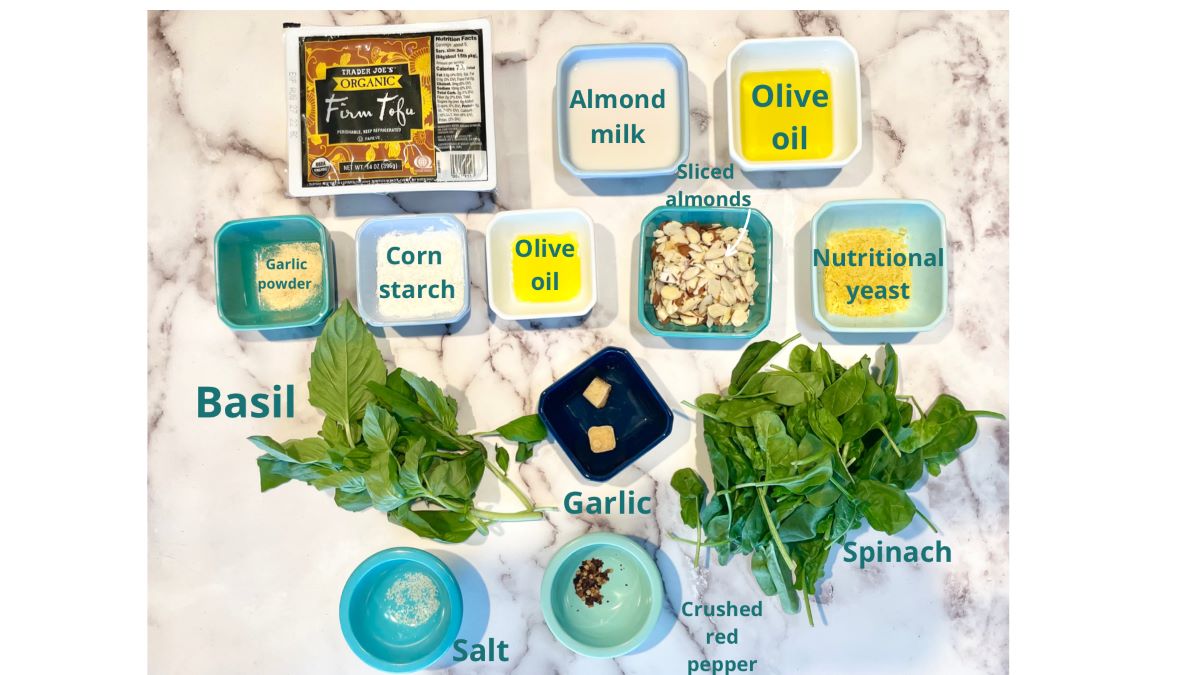 Pesto tofu ingredients in small colorful dishes atop marble. Ingredients include: firm tofu, almond milk, garlic powder, corn starch, olive oil, sliced almonds, nutritional yeast, basil leaves, garlic, baby spinach leaves, salt, and crushed red pepper