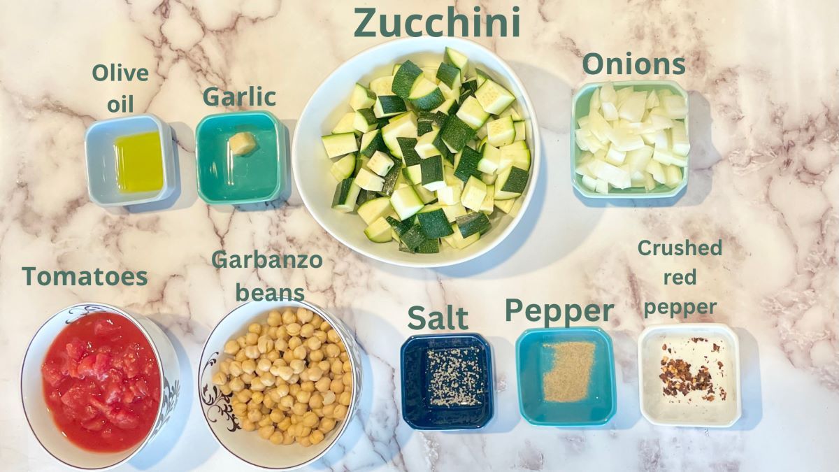 Zucchini stew ingredients atop a marble countertop. Ingredients are olive oil, garlic, zucchini, onions, diced canned tomatoes in juice, garbanzo beans, salt, pepper, and crushed red pepper.