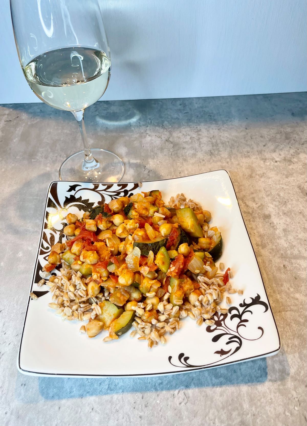 Zucchini stew served over farro atop a white plate with a swirl and next to a glass of white wine.