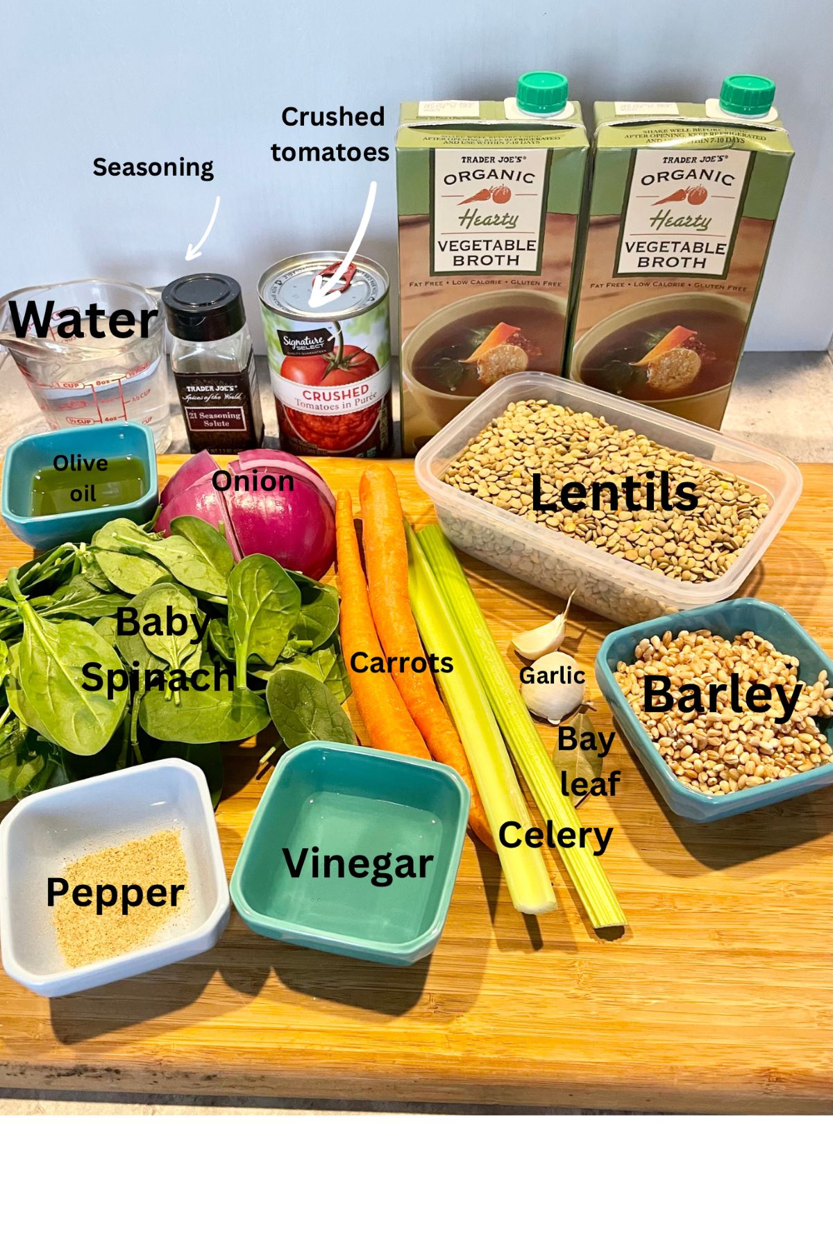 The picture displays all the ingredients needs to make carrot and lentil soup atop a wooden cutting board.  Ingredients pictured include: one cup of water, Trader Joe's 21 seasoning salute, one can of crushed tomatoes, 2 cartons of vegetable broth, olive oil, half a red onion, dried lentils, baby spinach leaves, 2 large carrots, 2 stalks of celery, 2 cloves of garlic, 1 bay leaf, dried barley, pepper, and white vinegar.
