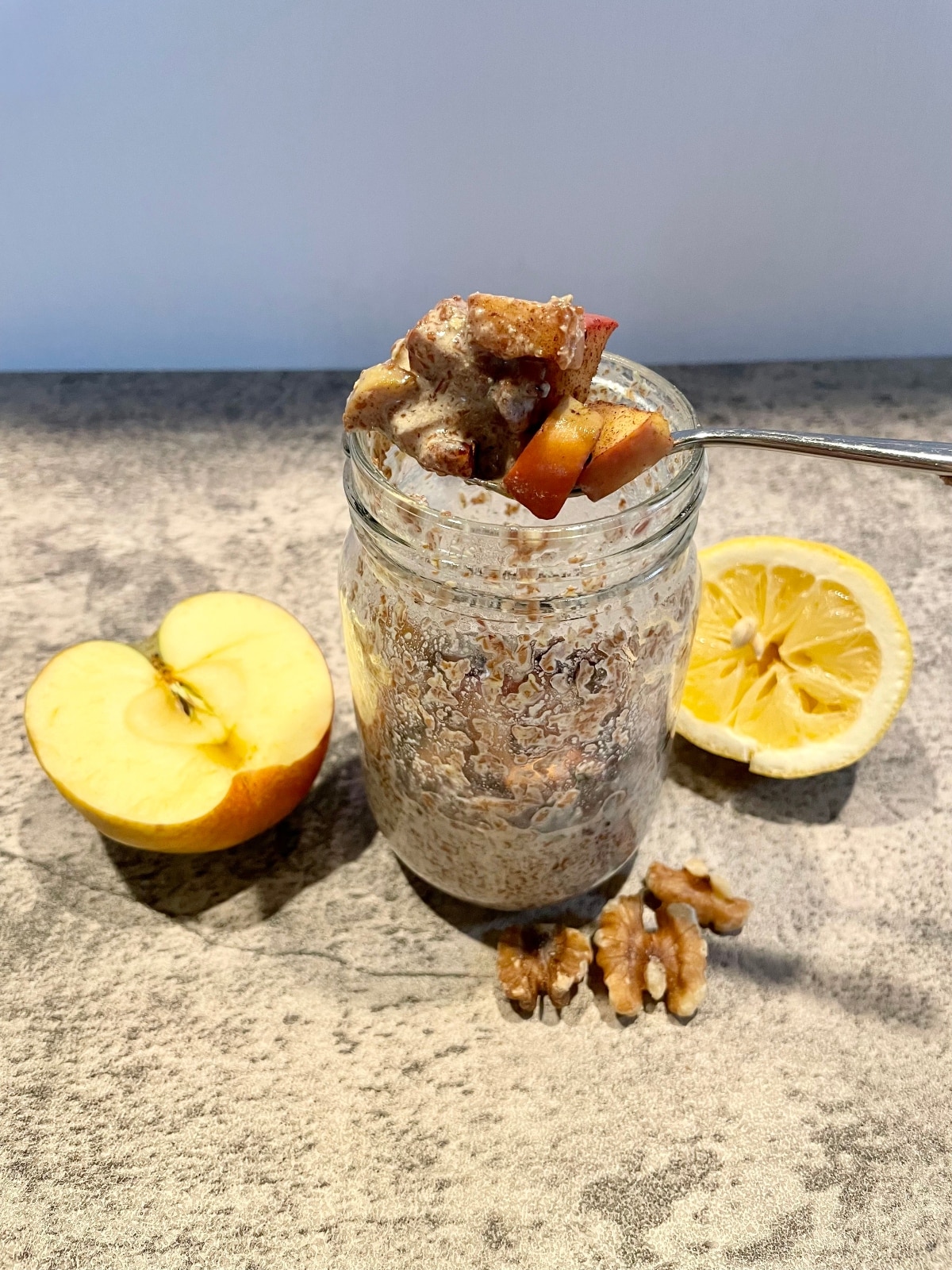 flaxseed pudding with apples, cinnamon, and walnuts in a mason jar atop a gray countertop.  Half a lemon, half an apple, and 3 walnuts halves are next to the jar.