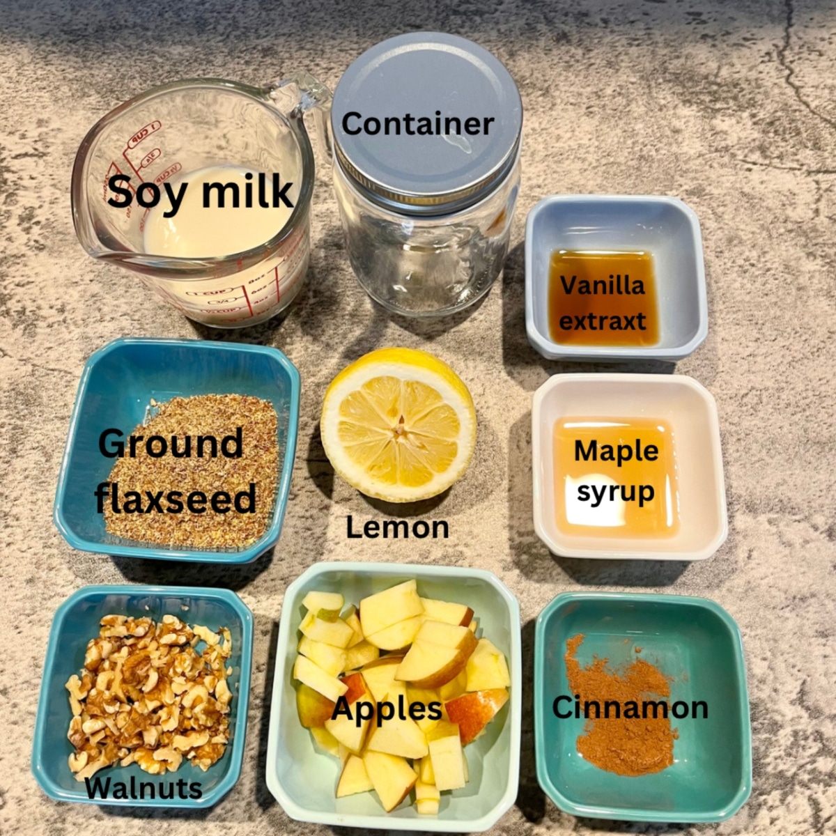 A picture showing what is needed for flaxseed pudding, which includes: soy milk, a container, vanilla extract, ground flaxseed, lemon, maple syrup, walnuts, apples, and cinnamon.  Each ingredients is in a small dish atop a gray countertop.