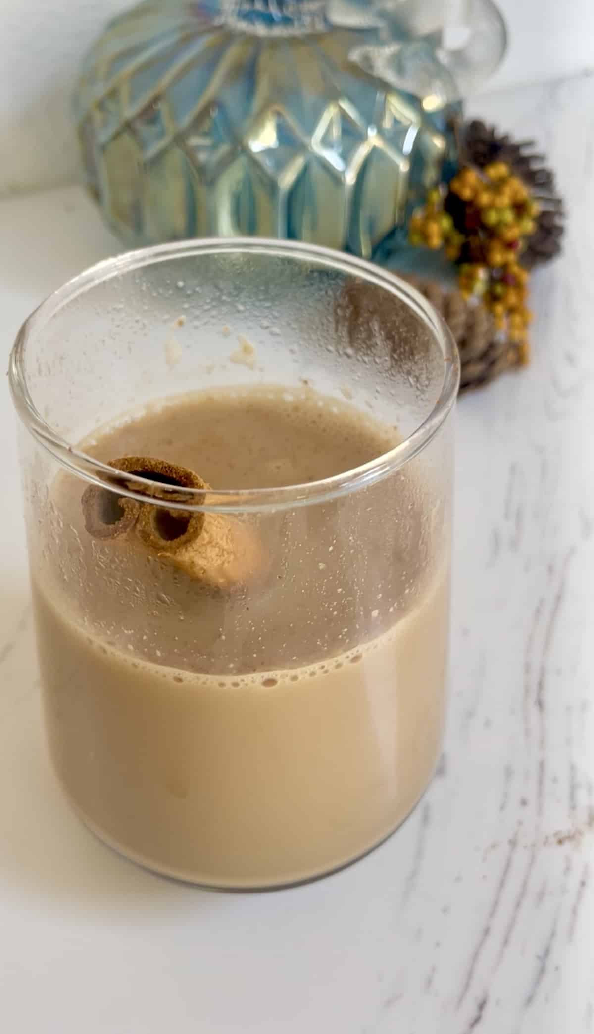 Pumpkin spice chai latte in a clear glass with a cinnamon stick atop a white countertop and in front of a blue glass pumpkin and other fall decorations.