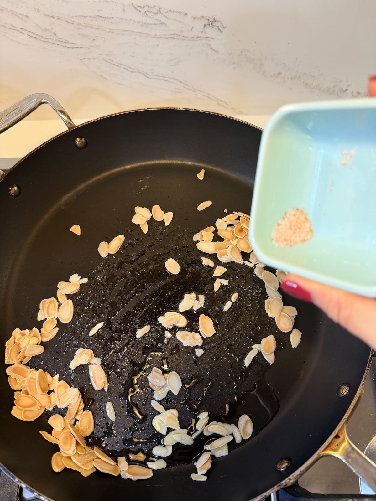 Pouring the salt from a light blue ramekin into the saucepan with olive oil and sliced almonds.