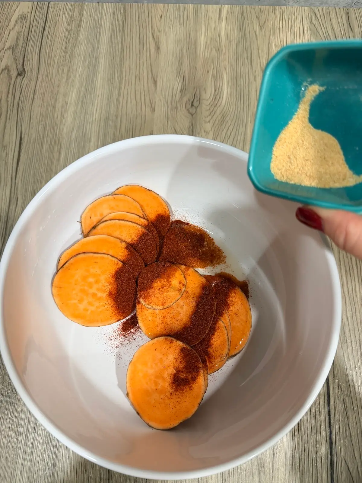 A large white bowl with sliced sweet potatoes and paprika atop a wooden surface. Above the bowl is a blue ramekin with garlic powder that is about to be added to the potatoes.