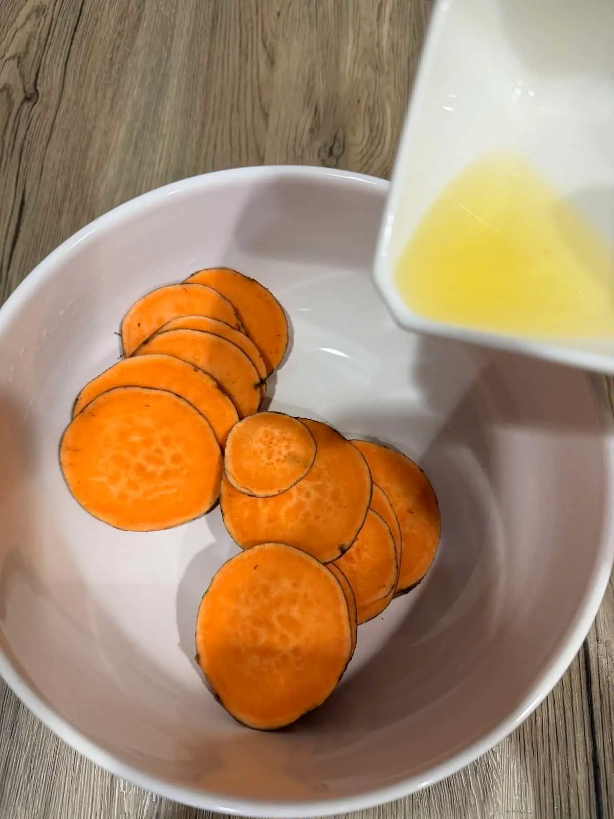 sliced sweet potatoes in a large white bowl atop a wooden surface. Above the bowl is a white ramekin with oil that is about to be added to the potatoes.