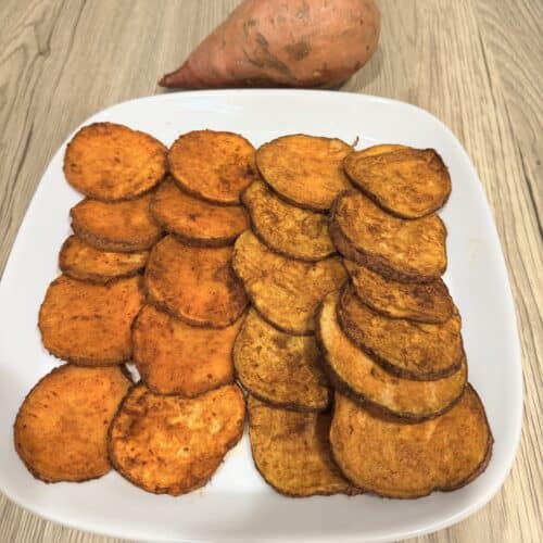 Air fryer sweet potato slices 2 ways on a white dish that is atop a wood surface. A whole sweet potato is pictured by the plate.