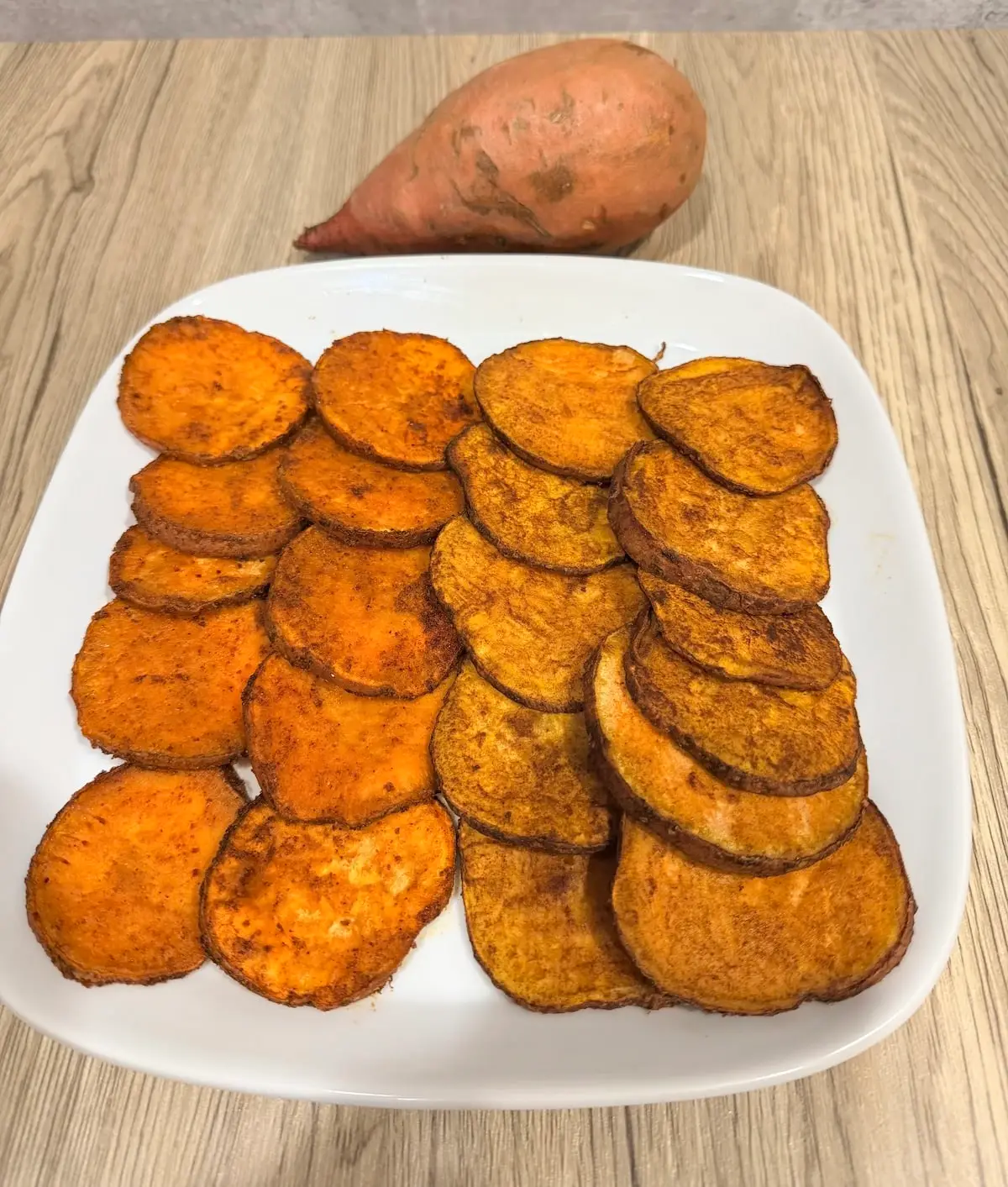 Air fryer sweet potato slices 2 ways on a white dish that is atop a wood surface. A whole sweet potato is pictured by the plate.