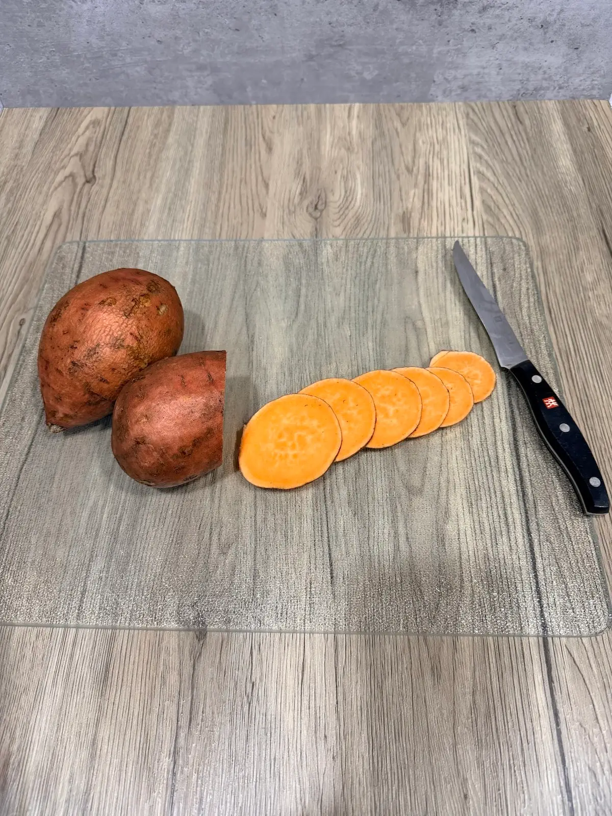 Whole sweet potatoes and thin slices of sweet potatoes atop and cutting board and wood surface.