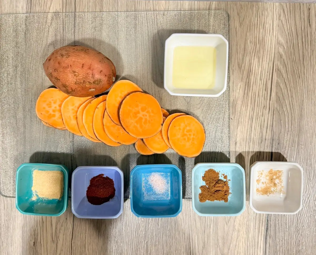 Atop a wood surface, a whole sweet potato, sweet potato slices, a white ramekin with oil, a blue ramekin with garlic powder, a blue ramekin with smoked paprika, a blue ramekin with salt, a blue ramekin with cinnamon, and a white ramekin with ginger powder.