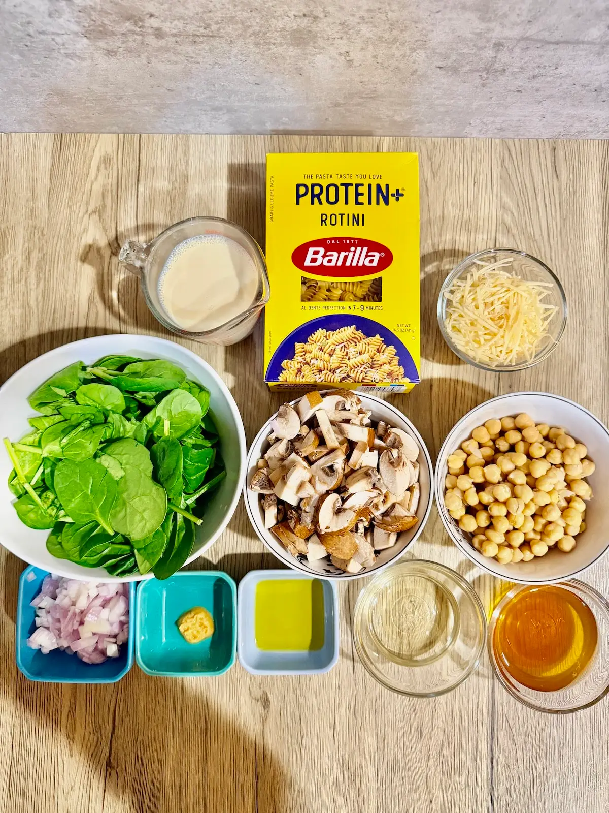 creamy vegetarian pasta ingredients atop a wooden surface- soy milk in a glass, protein pasta in the box, parmesan cheese in a glass bowl, spinach in a white bowl, chopped mushrooms in a white bowl, garbanzo beans in a white bowl, chopped shallots in a blue ramekin, crushed garlic in a blue ramekin, olive oil in a blue ramekin, white wine in a glass, and veggie broth in a glass.