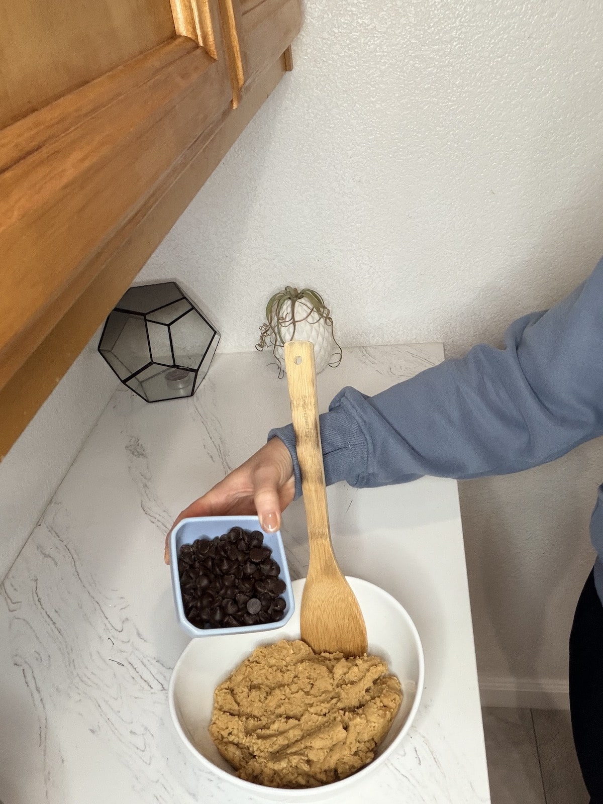 Chocolate chips in a blue ramekin. Cookie dough in a large white bowl and a mixing spoon.