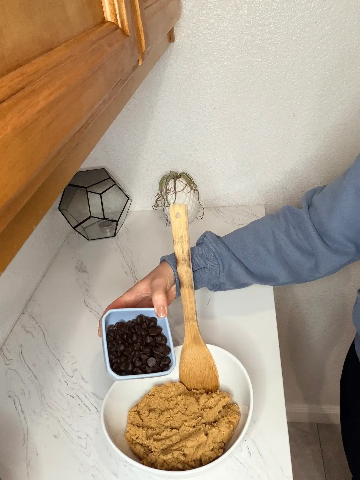 Chocolate chips in a blue ramekin. Cookie dough in a large white bowl and a mixing spoon.