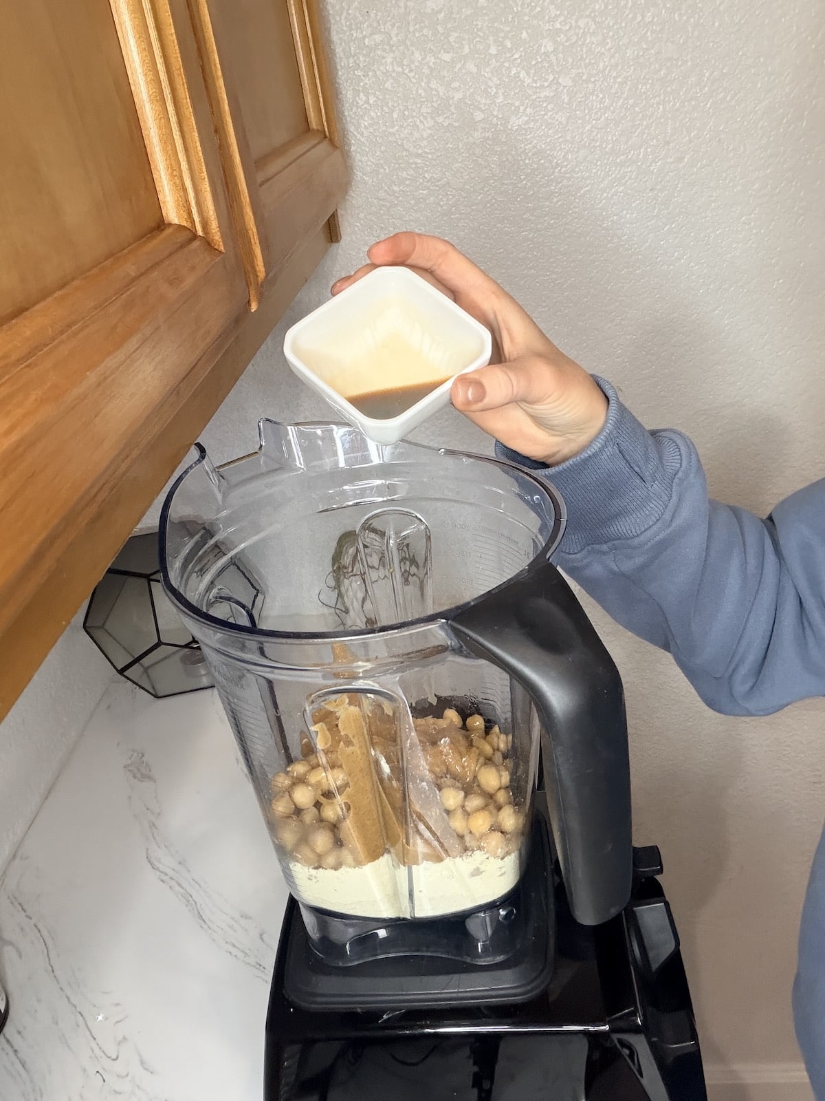 adding maple syrup to a blender container with chickpeas, almond flour, protein powder, vanilla extract, and peanut butter.