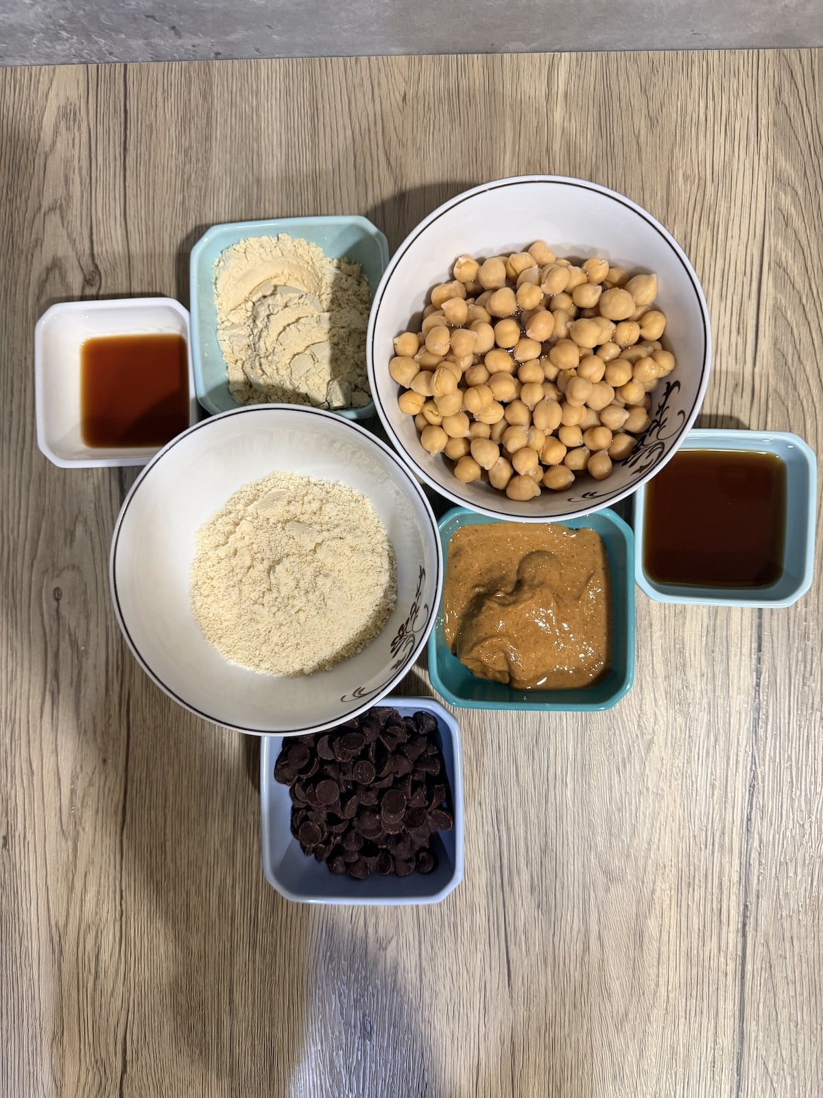 A white bowl with chickpeas, blue ramekin with maple syrup, a blue ramekin with peanut butter, a blue ramekin with chocolate chips, a white bowl with almond flour, a white ramekin with vanilla extract, and a blue ramekin with protein powder atop a wooden surface.