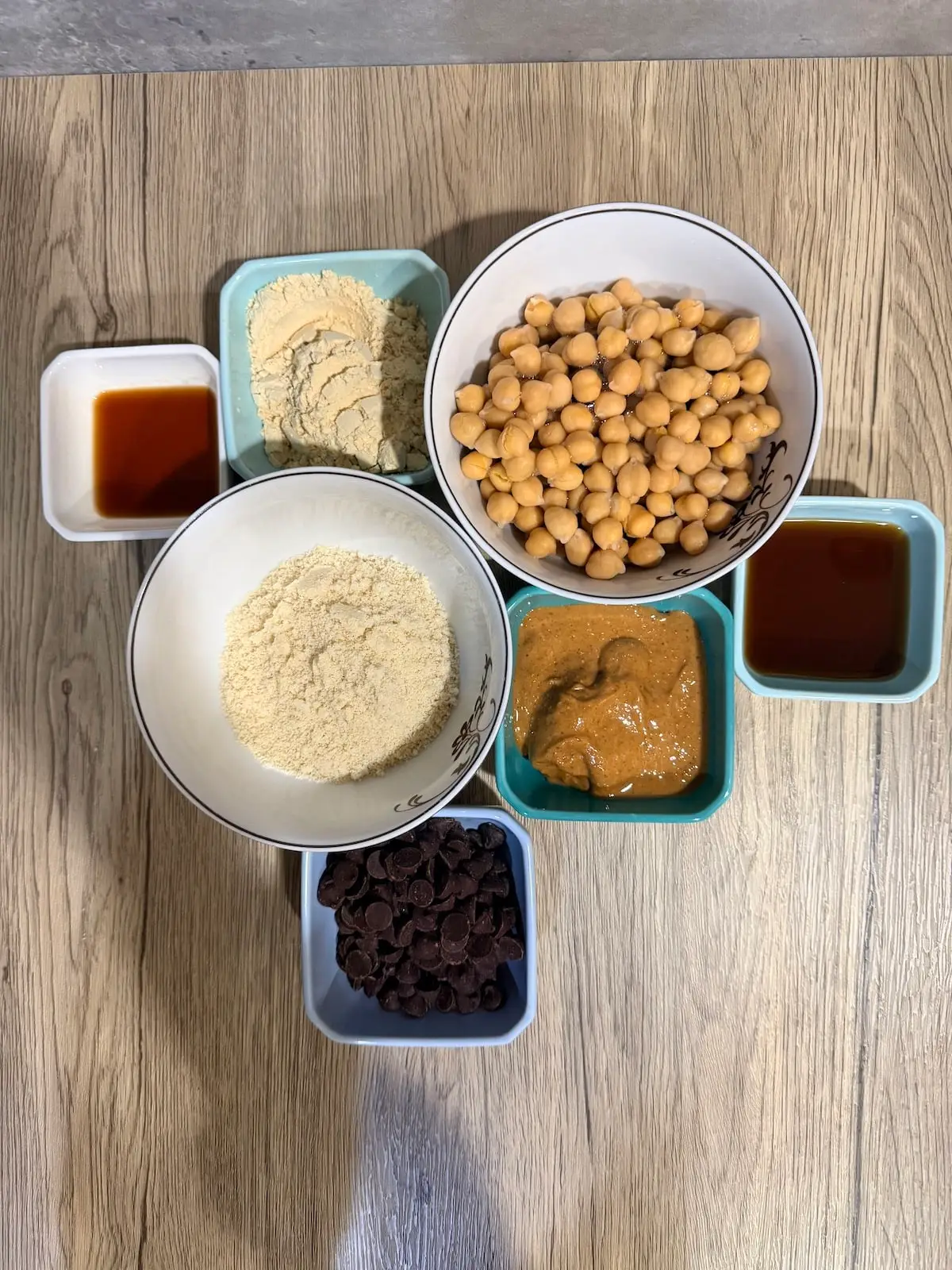 A white bowl with chickpeas, blue ramekin with maple syrup, a blue ramekin with peanut butter, a blue ramekin with chocolate chips, a white bowl with almond flour, a white ramekin with vanilla extract, and a blue ramekin with protein powder atop a wooden surface.
