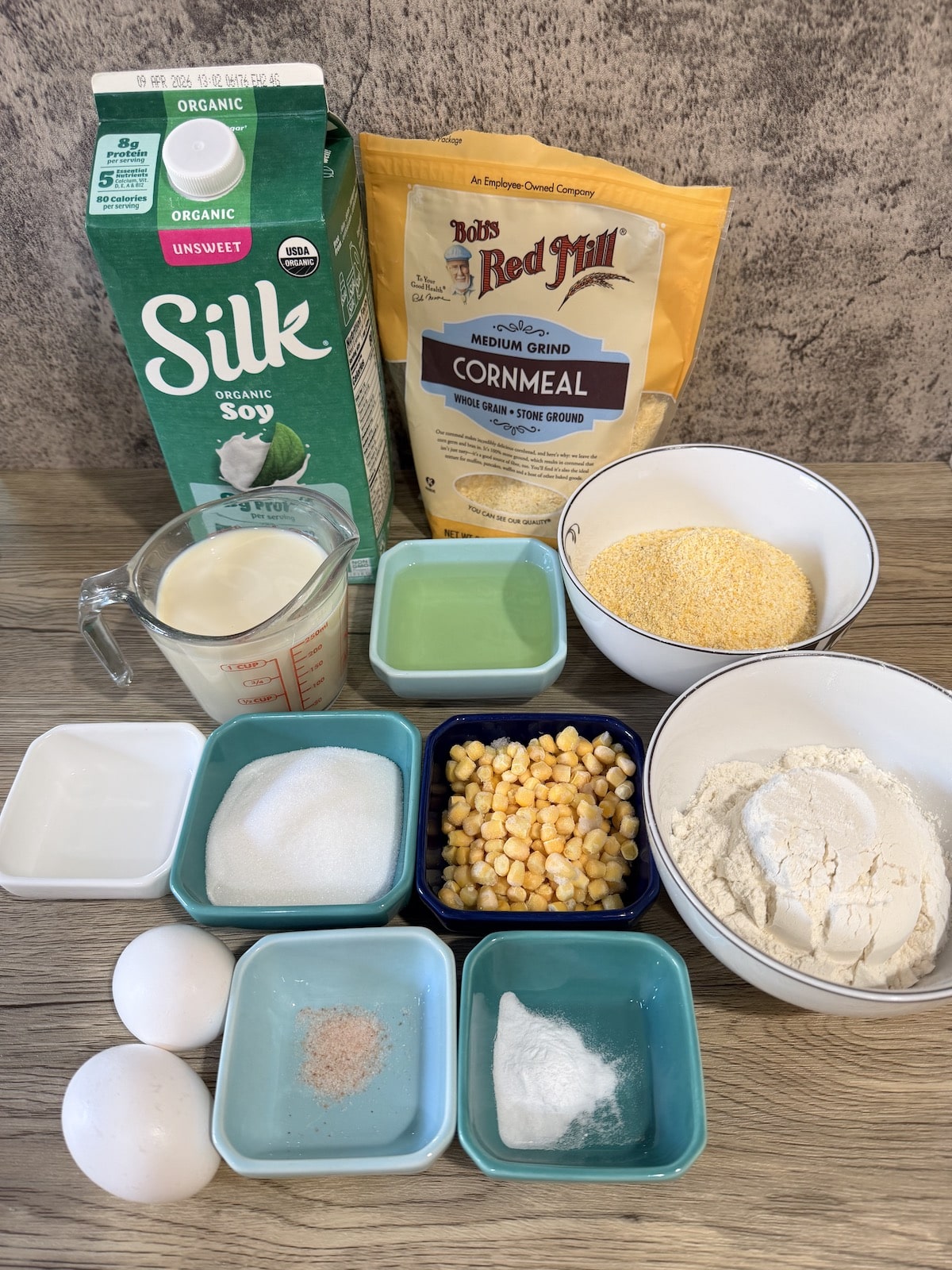 A carton of Silk organic unsweeted soy milk, bag of Bob's Redmill cornmeal, measuring cup with soy milk, blue ramekin with oil, white bowl with cornmeal, whte ramekin with vinegar, blue ramekin with sugar, blue ramekin with corn, white bowl with flour, 2 eggs, blue ramekin with salt, and blue ramekin with baking soda atop a wooden surface.