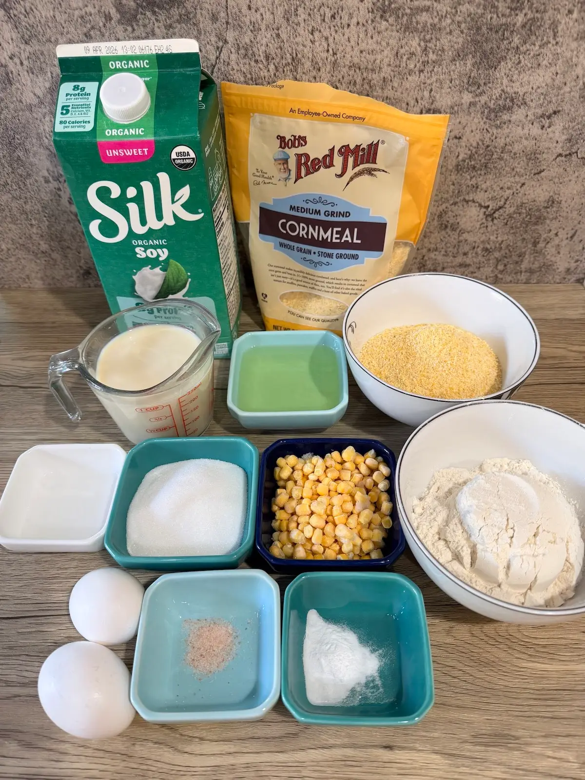 A carton of Silk organic unsweeted soy milk, bag of Bob's Redmill cornmeal, measuring cup with soy milk, blue ramekin with oil, white bowl with cornmeal, whte ramekin with vinegar, blue ramekin with sugar, blue ramekin with corn, white bowl with flour, 2 eggs, blue ramekin with salt, and blue ramekin with baking soda atop a wooden surface.