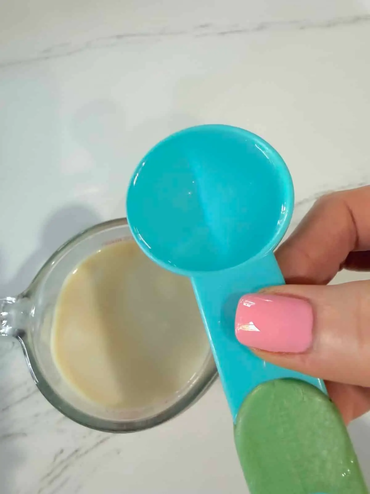 A blue tablepoon with white vinegar hovering over a measuring cup with unsweetened soy milk.