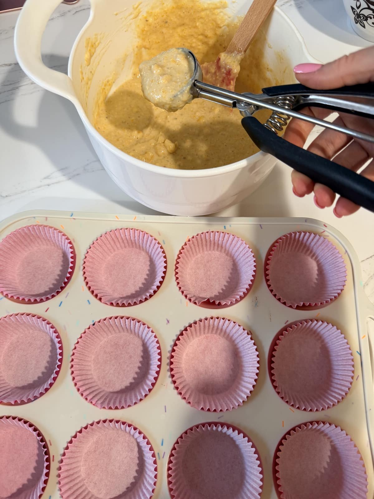 A white bowl with buttermilk cornbread muffin batter and cookie scoop holding the batter. A white muffin tin lined with red cupcake liners.