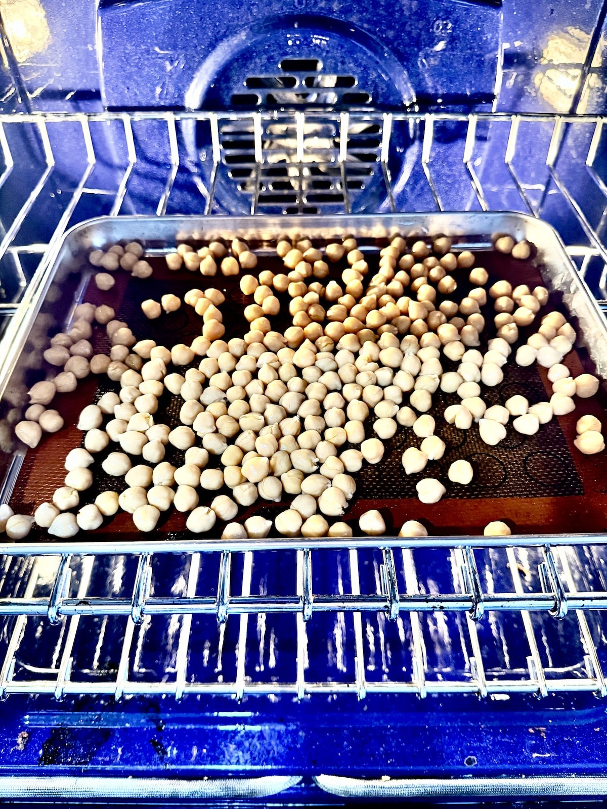 garbanzo beans atop a silicon lined baking sheet on the top rack in an oven that is blue on the inside.