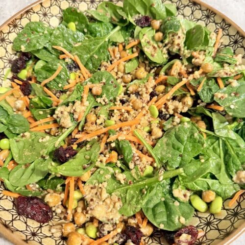 A large bowl with spinach leaves, shredded carrots, quinoa, garbanzo beans, edamame, dried cranberries, and tahini salad dressing.