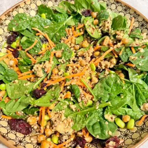 A large bowl with spinach leaves, shredded carrots, quinoa, garbanzo beans, edamame, dried cranberries, and tahini salad dressing.