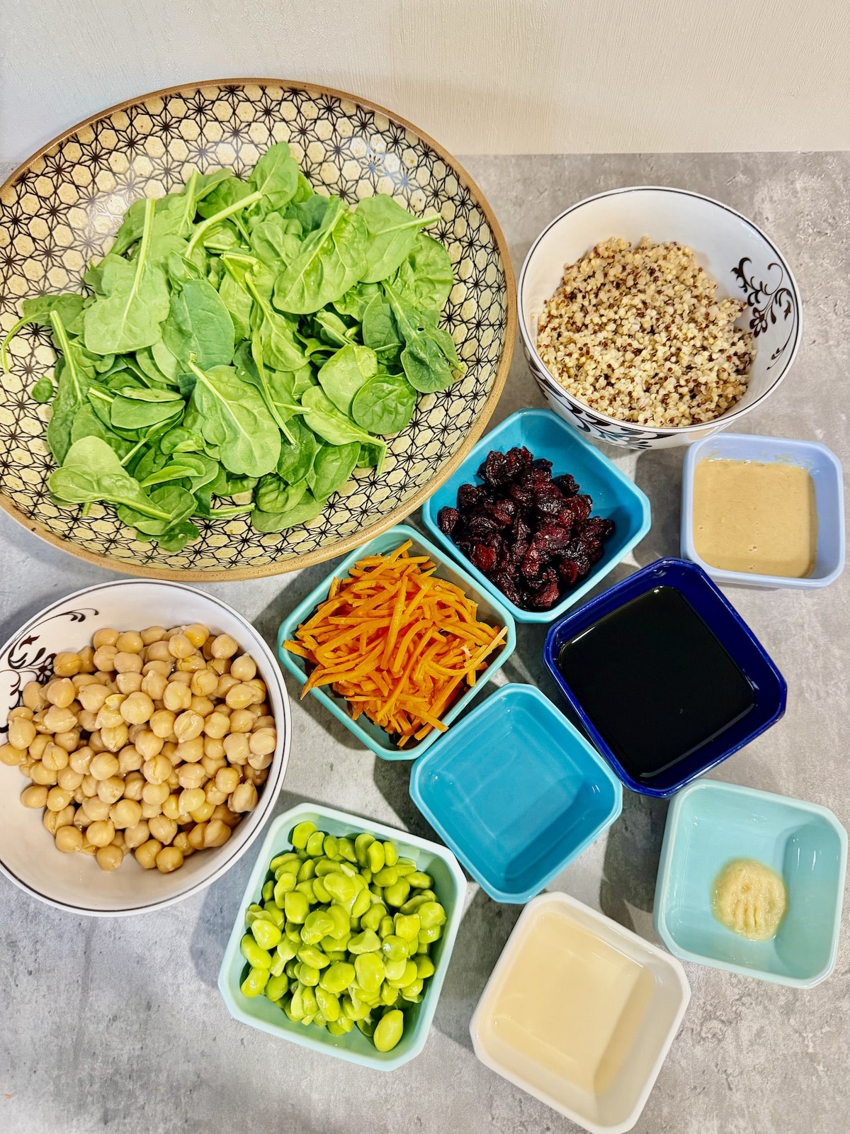 large bowl with baby spinach leaves, small white bowl with garbanzo beans, blue ramekin with shelled edamame, blue ramekin with water, blue ramekin with shredded carrots, blue ramekin with dried cranberries, blue ramekin with soy sauce, blue ramekin with crushed garlic, white ramekin with rice vinegar, blue ramekin with tahini, and white bowl with cooked tri-color quinoa.