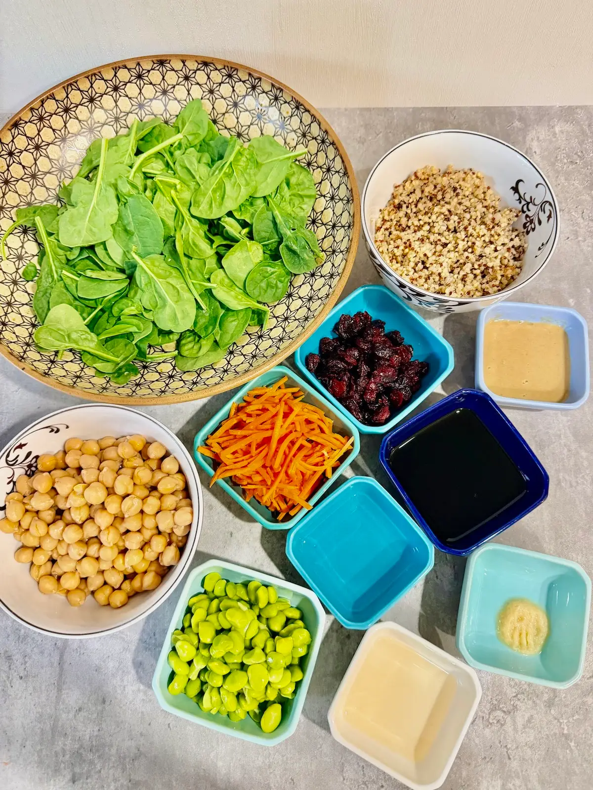 large bowl with baby spinach leaves, small white bowl with garbanzo beans, blue ramekin with shelled edamame, blue ramekin with water, blue ramekin with shredded carrots, blue ramekin with dried cranberries, blue ramekin with soy sauce, blue ramekin with crushed garlic, white ramekin with rice vinegar, blue ramekin with tahini, and white bowl with cooked tri-color quinoa.