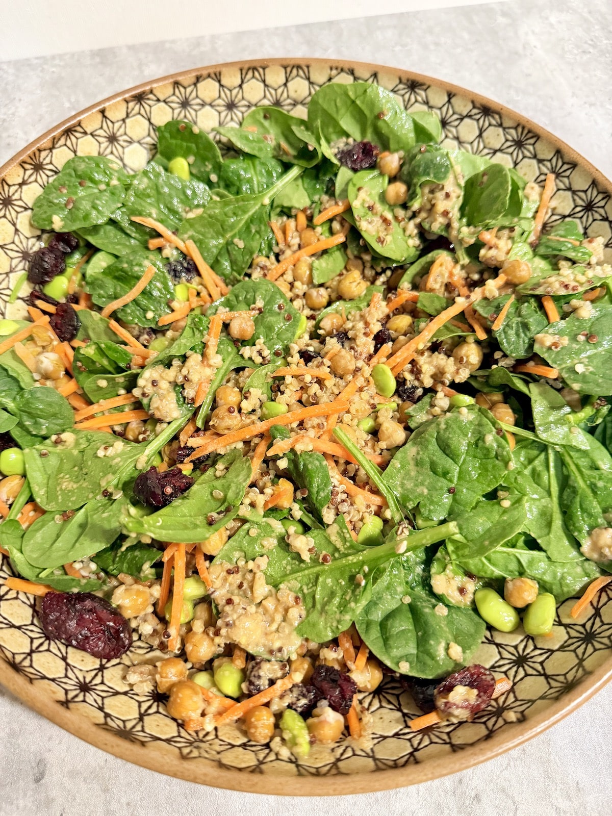 A large bowl with spinach leaves, shredded carrots, quinoa, garbanzo beans, edamame, dried cranberries, and tahini salad dressing.