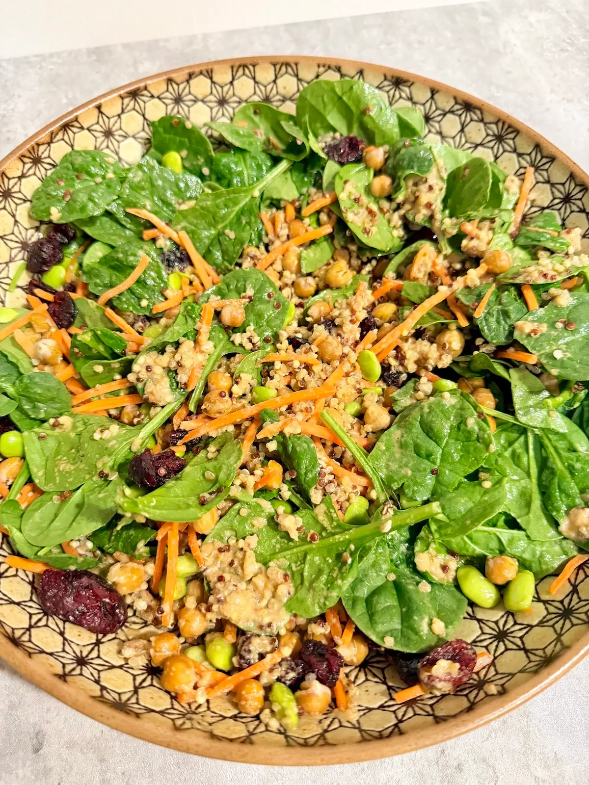 A large bowl with spinach leaves, shredded carrots, quinoa, garbanzo beans, edamame, dried cranberries, and tahini salad dressing.