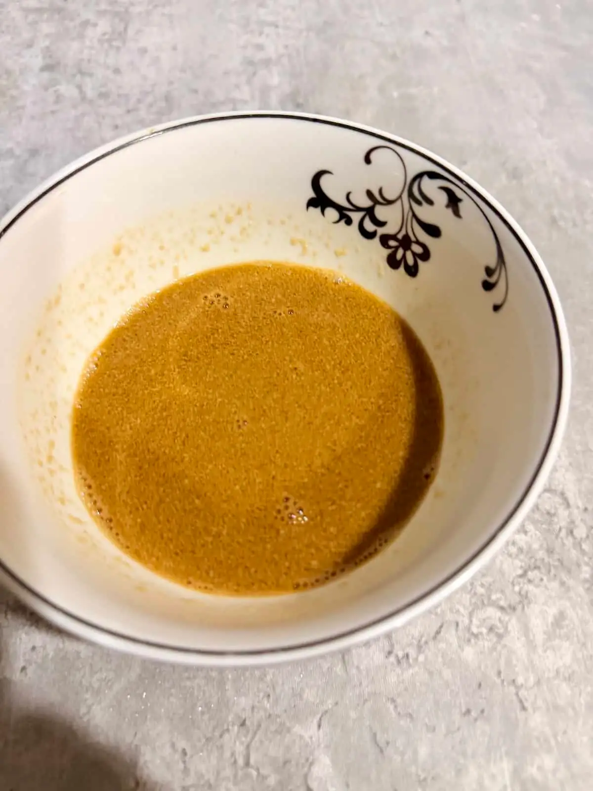 Tahini dressing in a white bowl with a black swirl pattern atop a gray surface.