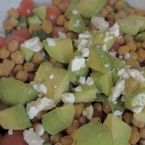 An up close shot of a bowl filled with roasted chickpeas, sliced cherry tomatoes, diced cucumber, chopped avocado, greens, and feta cheese.