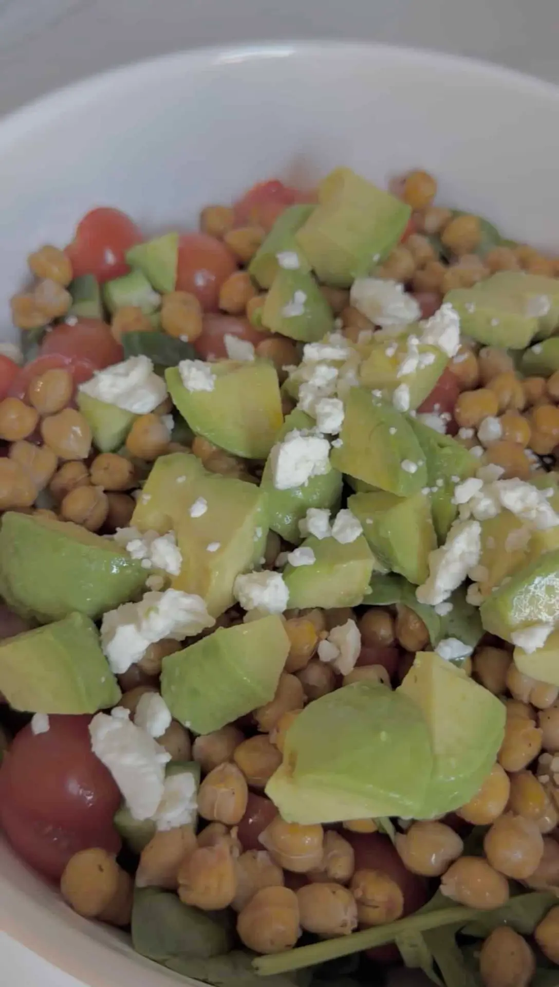 An up close shot of a bowl filled with roasted chickpeas, sliced cherry tomatoes, diced cucumber, chopped avocado, greens, and feta cheese.