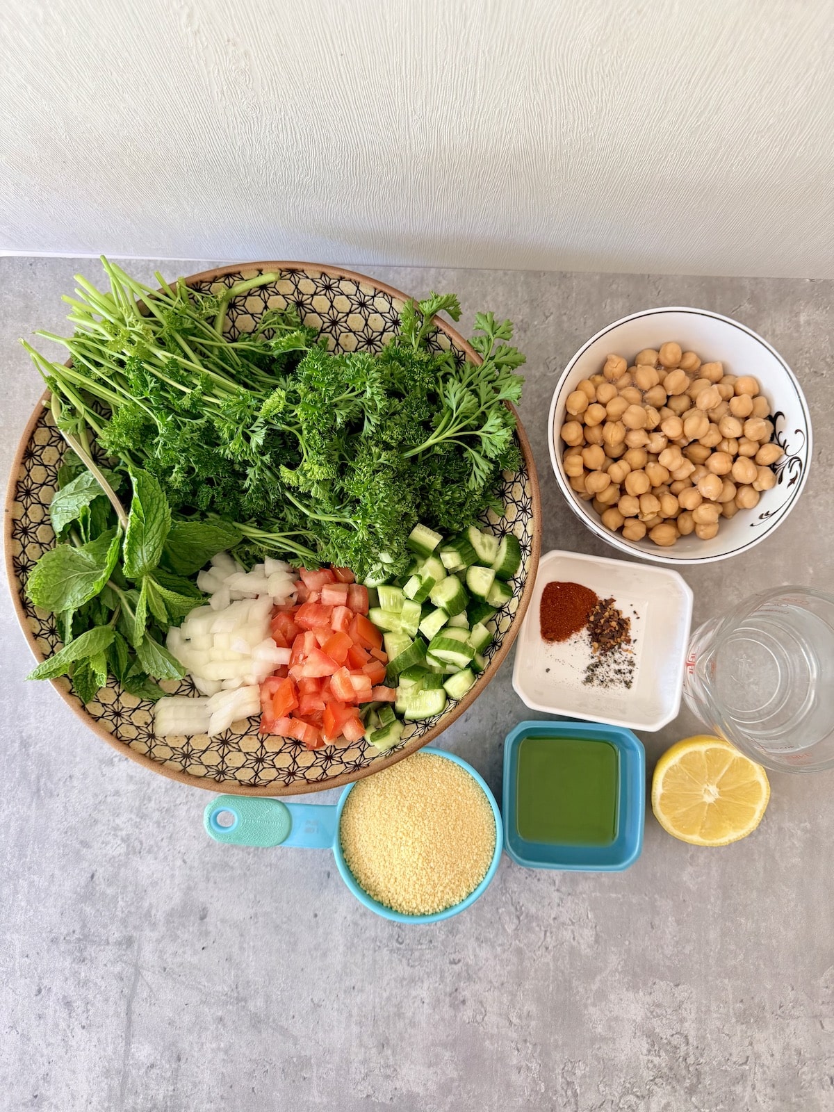 A large patterned bowl with parsley, mint leaves, diced onion, chopped tomato, and chopped cucumber aside a white bowl with chickpeas, a white ramiken with spices, half a lemon, a cup of dry couscous, a cup of water, and a white ramiken with olive oil. All atop a gray surface.