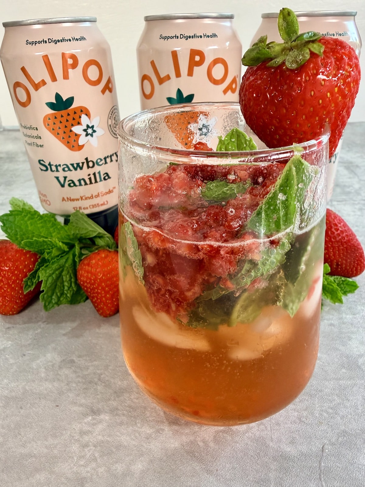 Three cans of Strawberry Vanilla Olipop positioned behind a clear glass with a summer mocktail recipe made with ice, crushed berries, mint leaves, and Olipop Strawberry Vanilla. A strawberry garnishes the glass. Strawberries and mint leaves garnish the base of the cans.