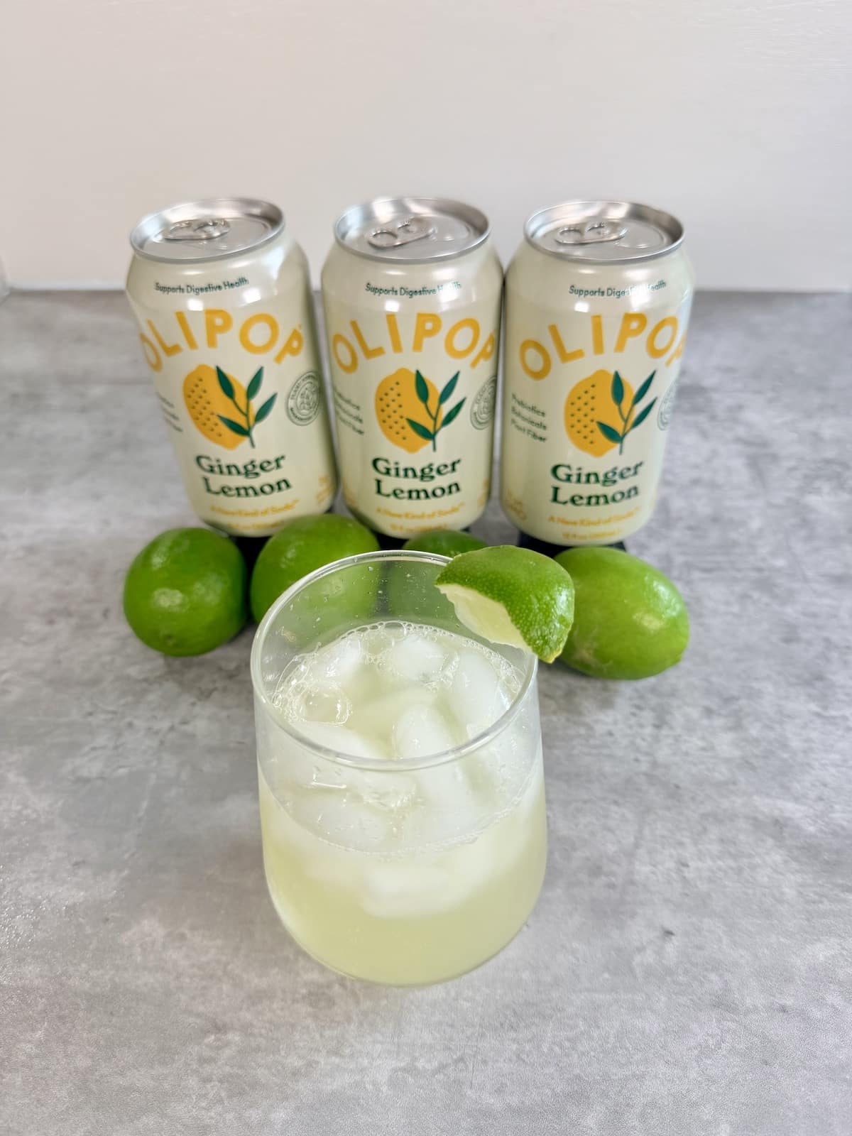 Three cans of Olipop Ginger Lemon positioned behind a summer mocktail recipe with lime juice, ice, and Olipop Ginger Lemon. Limes boarder the base of the cans.