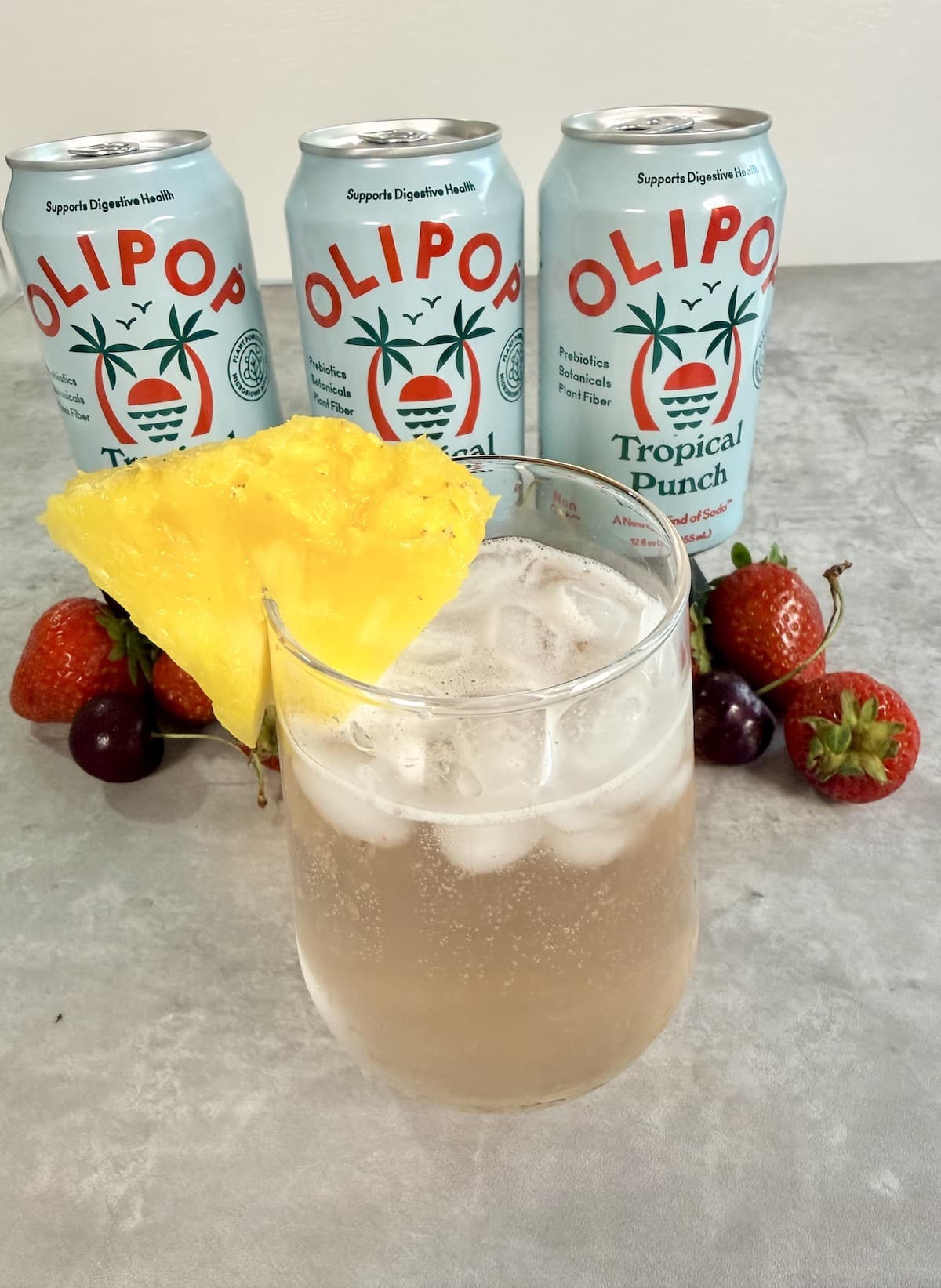 Three cans of Olipop Tropical Punch positioned behind a clear glass with a bubbly, pink summer mocktail recipe with a pineapple wedge garnish. Decorative strawberries and cherries are at the base of the cans.