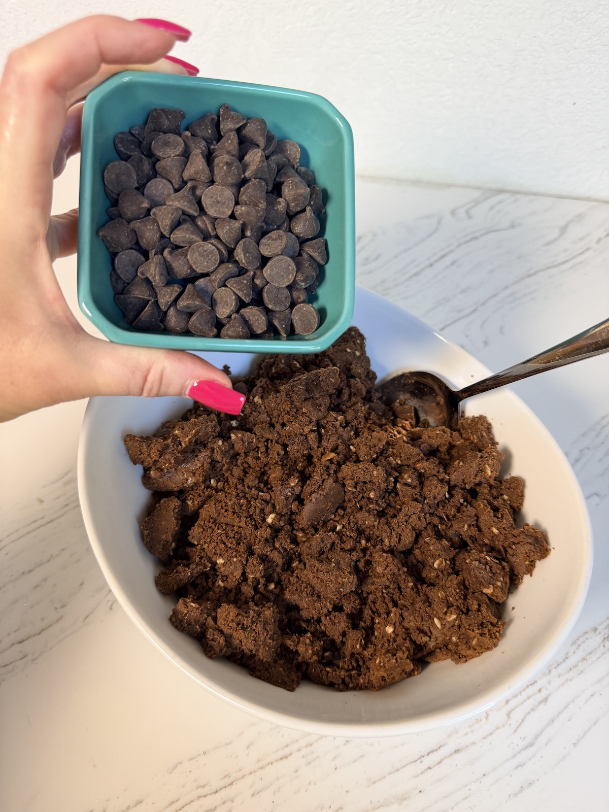 Date Protein Ball with Peanut Butter & Chocolate batter in a white bowl with teal ramiken of chocolate chips held above it.