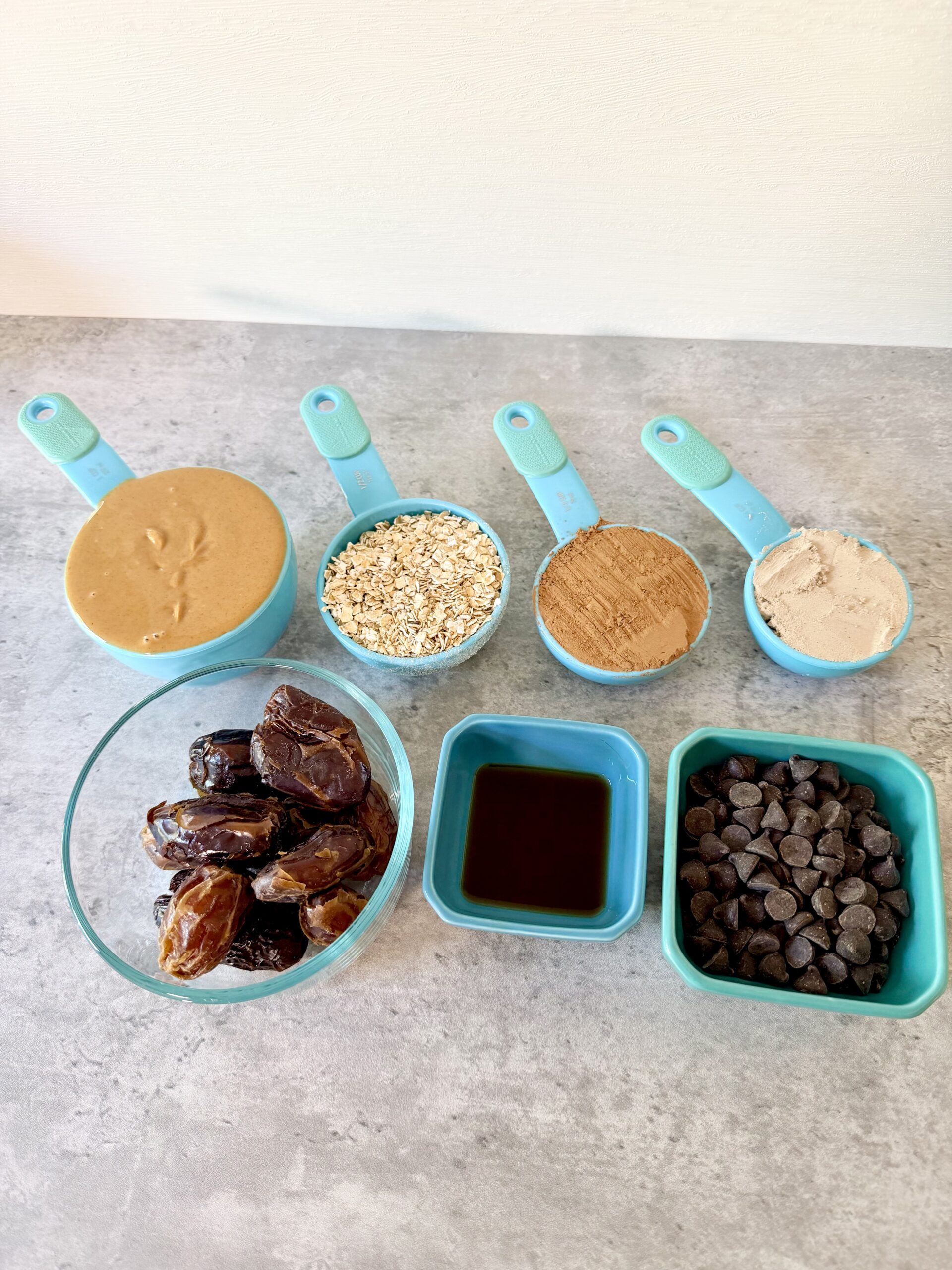 Teal measuring cups with peanut butter, rolled oats, cocoa powder, chocolate protein powder, a clear bowl with Medjool dates, a teal ramiken with vanilla extract, and a teal ramiken with chocolate chips atop a gray surface.