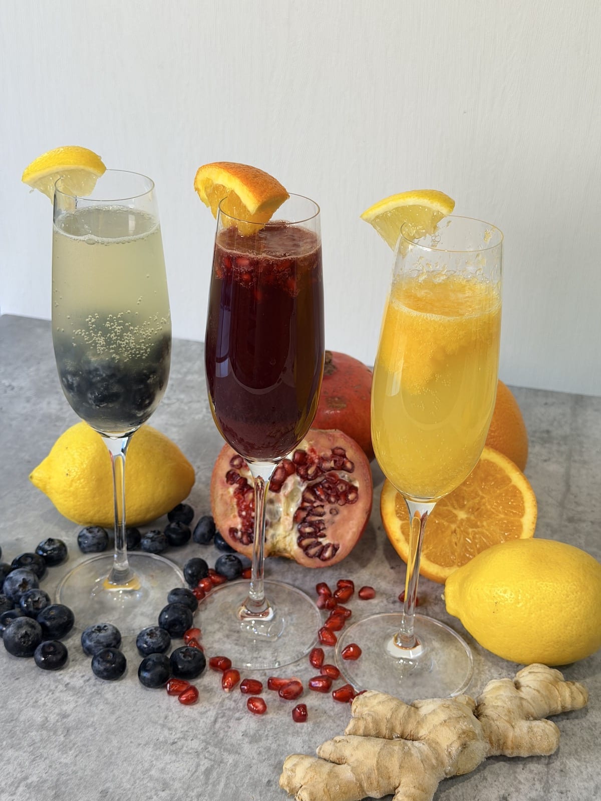 Champagne flutes with blueberry lemon fizz, pomegranate citrus spritz, and peach ginger bellini atop a grap surface. Lemons, pomegranates, pomegranate arils, blueberries, oranges, and ginger root decorate the surface.