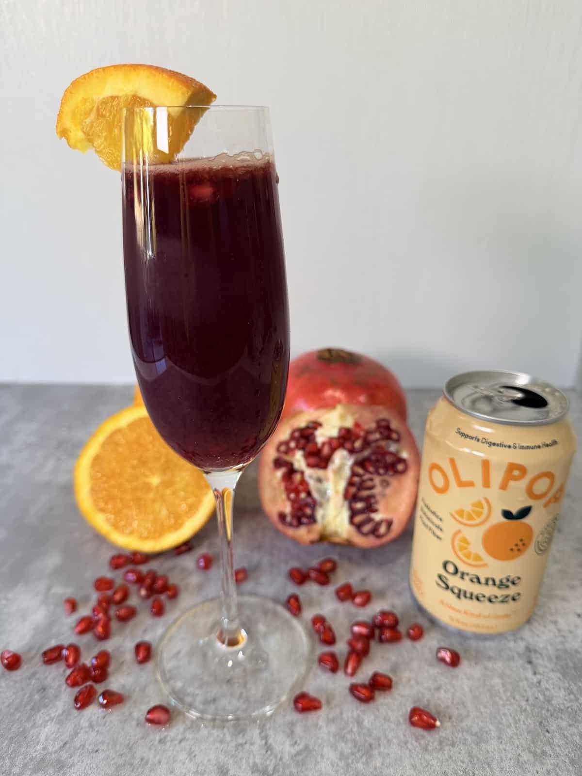 A champagne flute garnished with an orange slice and filled with orange juice, pomegranate juice, pomegranate arils, and Olipop Orange Squeeze atop a gray surface. An orange, pomegranate, and pomegranate arils decorate the surface. A can of Olipop Orange Squeeze is next to the champagne flute.