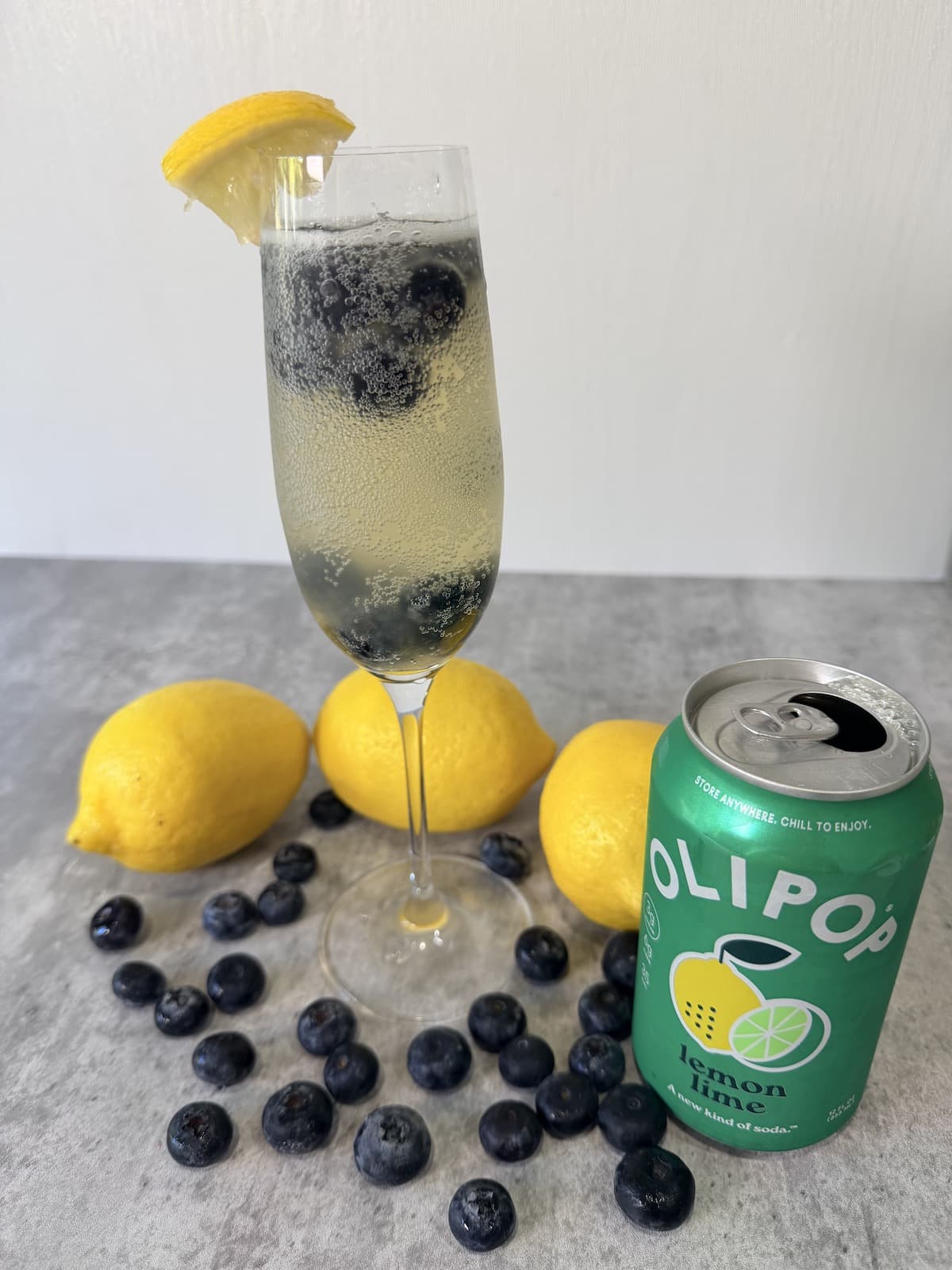 A champagne flute garnished with a lemon slice and filled with lemon juice, blueberries, and Olipop Ginger Lemon atop a gray surface. Lemons and blueberries surround the champagne flute. A can of Olipop Lemon Lime is next to the champagne flute.