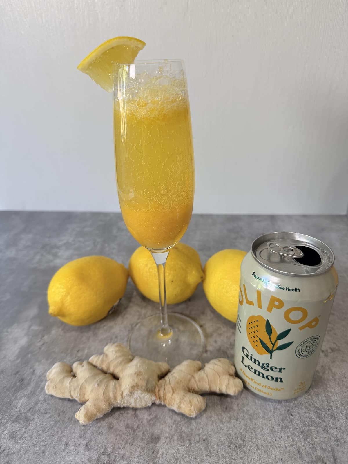 Champagne flute garnish with a lemon wedge and filled with peach puree, lemon juice, and Olipop Ginger Lemon atop a gray surface. 3 lemons  are behind the champagne flute. A can of Olipop Ginger Lemon is next to the champagne flute. A large piece of ginger root is infront of the glass.