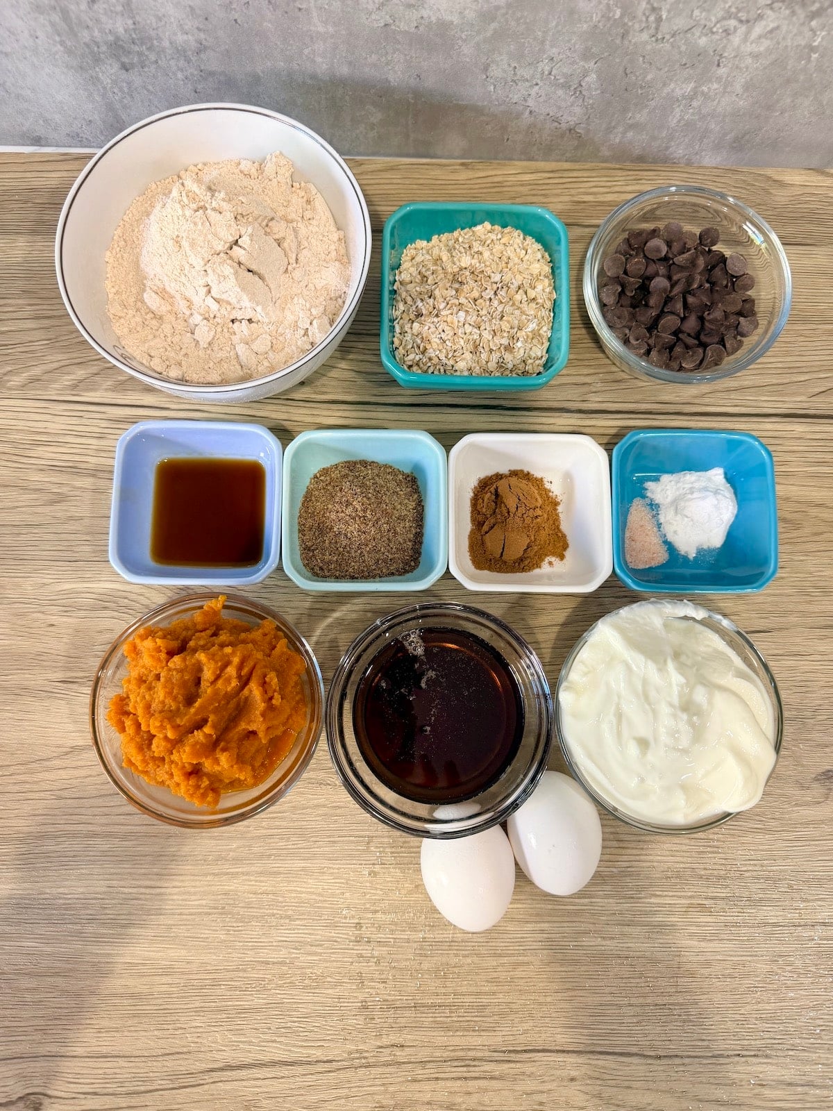 A white bowl with whole wheat flour, blue ramekin with oats, a clear bowl with chocolate chips, a blue ramekin with vanilla extract, a blue ramekin with ground flaxseed,  a white ramekin with spices, a blue ramekin with salt and baking soda, a clear bowl with pumpkin puree, a clear bowl with maple syrup, a clear bowl with Greek yogurt and 2 eggs atop a wooden surface.